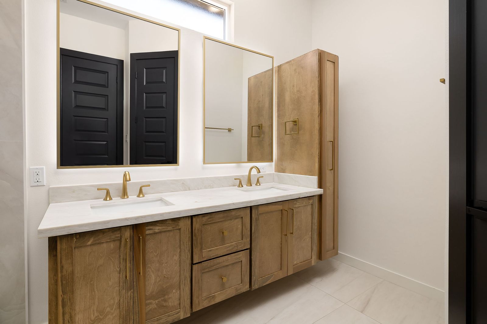 Primary bathroom with a wood double vanity and brushed-gold fixtures — The Black & Stone, a Kratos Construction custom home
